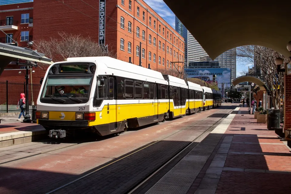 White and yellow DART light rail train in Dallas, Texas with brick building in background.